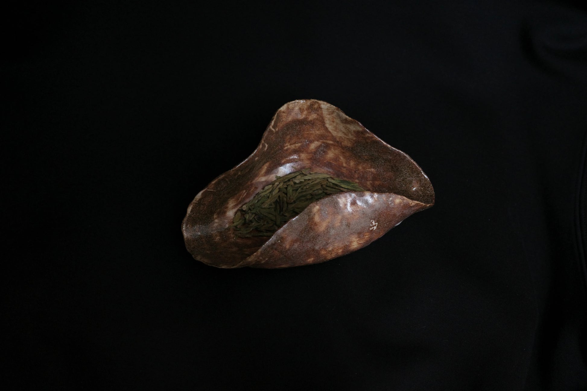 Handcrafted ceramic bowl with organic shape holding loose green tea leaves on black background