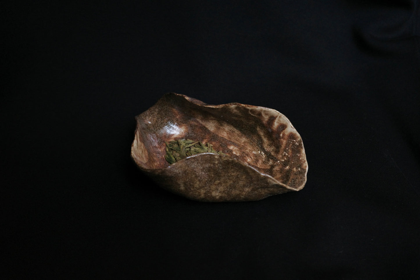 Handcrafted ceramic bowl with rustic brown glaze holding dried leaves on a dark background