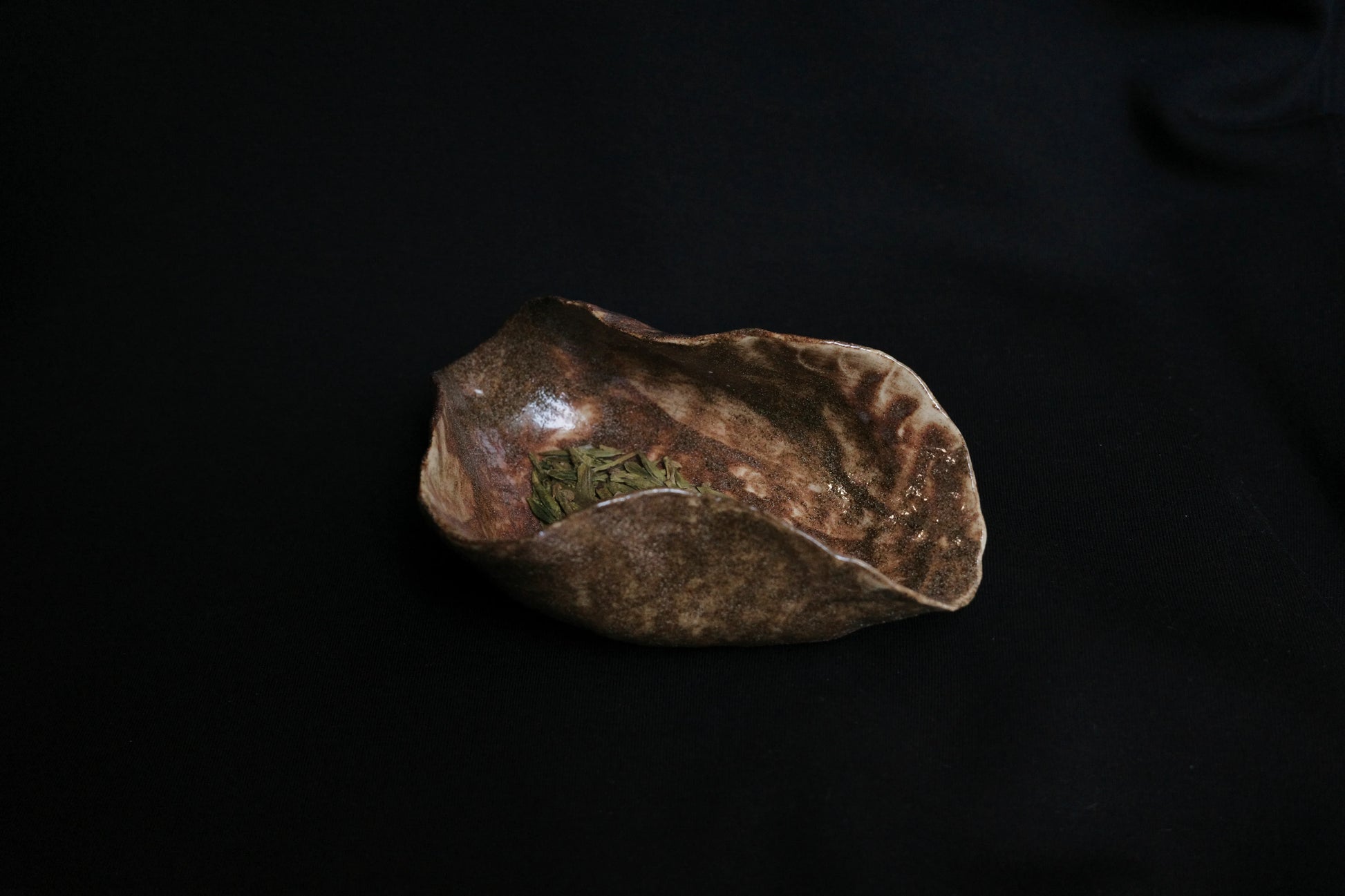 Handcrafted ceramic bowl with rustic brown glaze holding dried leaves on a dark background