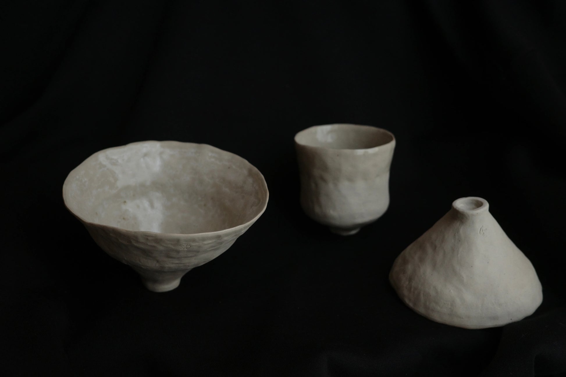 Minimalist handcrafted ceramic bowl, cup, and vase set on black fabric background